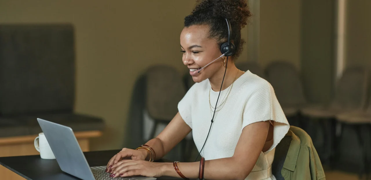 Das Bild zeigt eine lächelnde Kundenservice-Mitarbeiterin oder Telefonistin in einem modernen Büro. Sie trägt ein cremefarbenes kurzärmeliges Oberteil und ein Headset mit Mikrofon.
