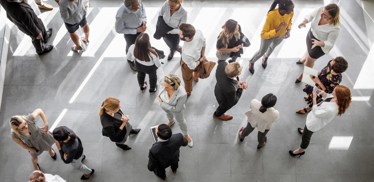 Das Bild zeigt eine Vogelperspektive einer Gruppe von etwa 20 Personen in Business- und Business-Casual-Kleidung, die auf einem modernen, hellgrauen Fliesenboden stehen.