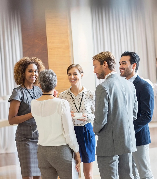 Das Bild zeigt eine professionelle Networking-Veranstaltung oder Business-Meeting in einem hellen, modernen Raum mit hohen Fenstern und bodenlangen, halbtransparenten Vorhängen. Im Vordergrund stehen fünf Personen in Business-Kleidung in einer lockeren Gesprächsrunde. Eine Frau mit lockigen Haaren in einem grauen Kleid unterhält sich mit einer Kollegin mit Kurzhaarschnitt und weißem Oberteil, die von hinten zu sehen ist. In der Mitte steht eine lächelnde Frau in gepunkteter Bluse und blauem Rock, die eine Kaffeetasse hält und ein Namensschild trägt. Rechts daneben stehen zwei Männer in hellgrauen Anzügen – einer von hinten, der andere im Profil sichtbar.