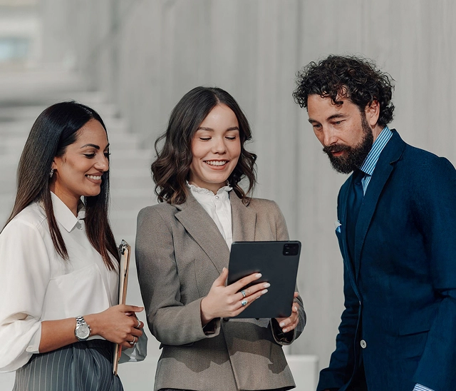 Das Bild zeigt drei Business-Professionals in formeller Kleidung, die gemeinsam auf ein Tablet schauen. In der Mitte steht eine lächelnde Frau in einem beigefarbenen Hosenanzug mit weißer Bluse, die das Tablet hält. Links neben ihr steht eine Kollegin in cremefarbener Bluse mit gestreifter Hose, die ein Clipboard in der Hand hält und eine Armbanduhr trägt. Rechts steht ein Mann mit Bart und dunklen lockigen Haaren in einem dunkelblauen Anzug mit hellblauem gestreiftem Hemd. Alle drei wirken konzentriert und interessiert an den Inhalten auf dem Bildschirm.